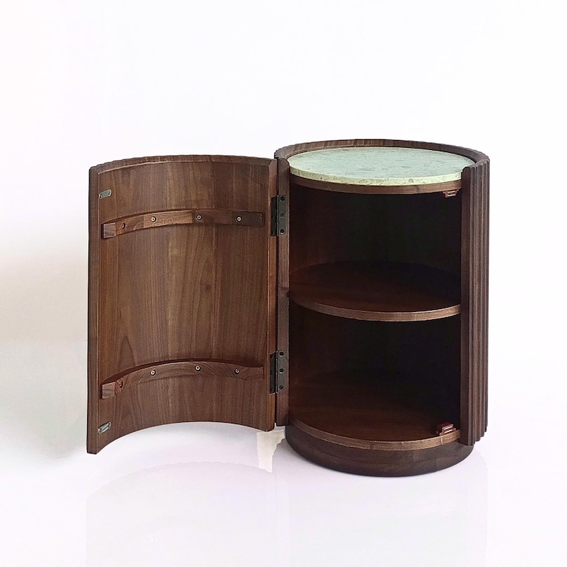 May include: A dark brown, cylindrical cabinet with an open door revealing two shelves. The top surface is a light green speckled stone. The cabinet has a curved door and a fluted exterior design.