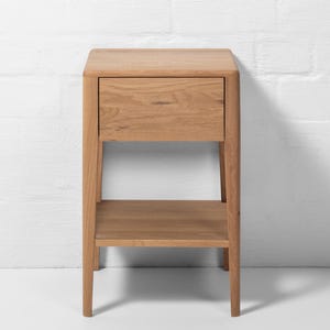 Front view of a handmade American white oak nightstand, crafted in a Scandinavian minimalist style. This small bedside table features one soft-close drawer with a smooth flat front and an open storage shelf below. Made from natural white oak hardwood, the piece stands on four tapered legs, highlighting clean lines, visible wood grain, and modern craftsmanship against a white brick wall.