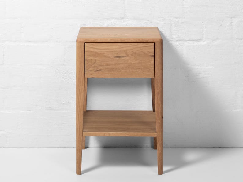 Front view of a handmade American white oak nightstand, crafted in a Scandinavian minimalist style. This small bedside table features one soft-close drawer with a smooth flat front and an open storage shelf below. Made from natural white oak hardwood, the piece stands on four tapered legs, highlighting clean lines, visible wood grain, and modern craftsmanship against a white brick wall.