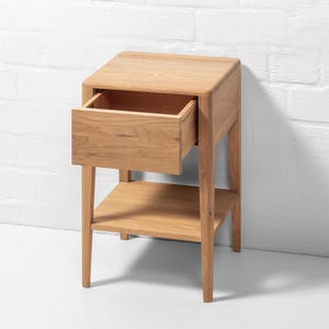 Front angle view of a handmade American white oak nightstand with a single drawer and open lower shelf. It features a Scandinavian-inspired minimalist design and natural wood grain texture. The drawer is shown open, revealing a matching oak interior. The open lower shelf provides extra storage or display space. Photographed against a plain white wall and floor, this solid oak bedside table showcases clean, straight lines and a warm natural wood tone, emphasizing its modern minimalist aesthetic.