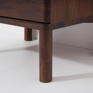 May include: Close-up of a dark brown wooden furniture piece with rounded corners and cylindrical legs. The wood grain is visible, and the legs are evenly spaced. The furniture is set against a white background.