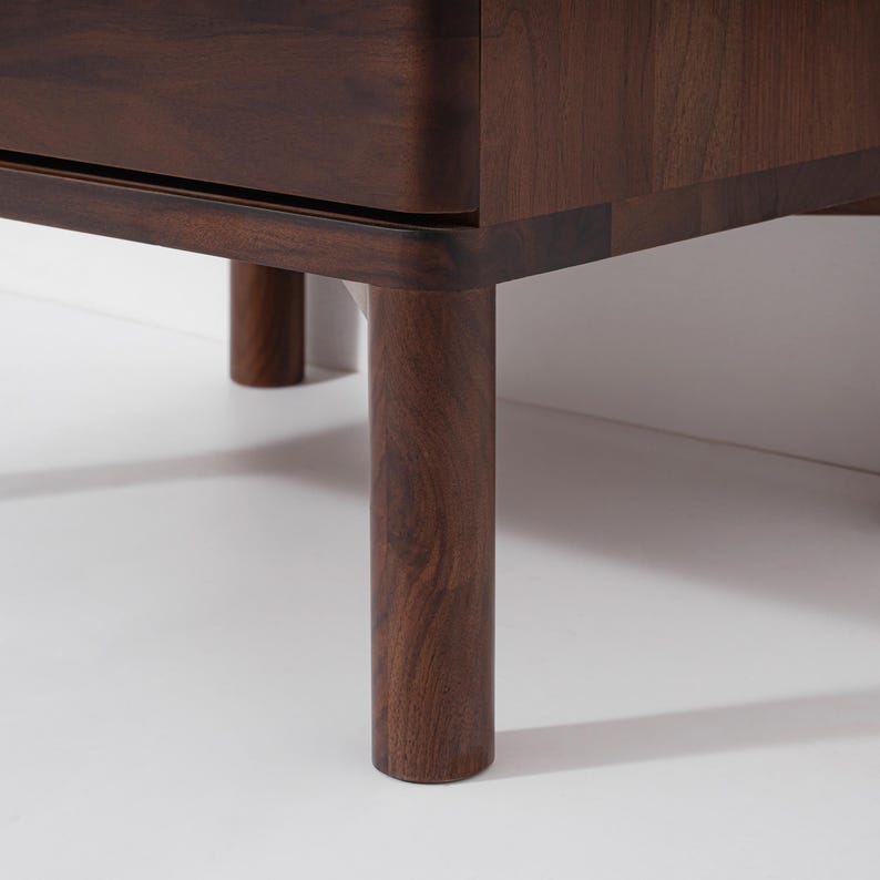 May include: Close-up of a dark brown wooden furniture piece with rounded corners and cylindrical legs. The wood grain is visible, and the legs are evenly spaced. The furniture is set against a white background.