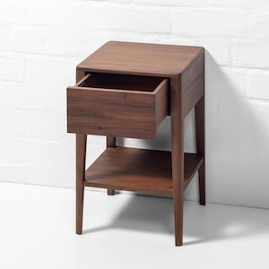 A solid walnut nightstand handcrafted from American walnut is shown from a front-right angle against a white brick wall. The wooden nightstand with drawer has the soft-close drawer pulled open, revealing an empty lower shelf and highlighting the rich walnut grain and quality craftsmanship. This compact bedside table exemplifies modern wood bedside furniture with its clean lines and sturdy build.