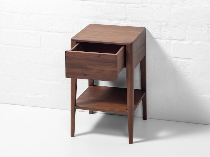 A solid walnut nightstand handcrafted from American walnut is shown from a front-right angle against a white brick wall. The wooden nightstand with drawer has the soft-close drawer pulled open, revealing an empty lower shelf and highlighting the rich walnut grain and quality craftsmanship. This compact bedside table exemplifies modern wood bedside furniture with its clean lines and sturdy build.