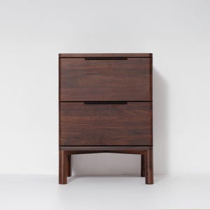 May include: A two-drawer wooden nightstand with a dark brown finish. The nightstand has a rectangular shape with rounded edges and black handles. It stands on four short, sturdy legs.