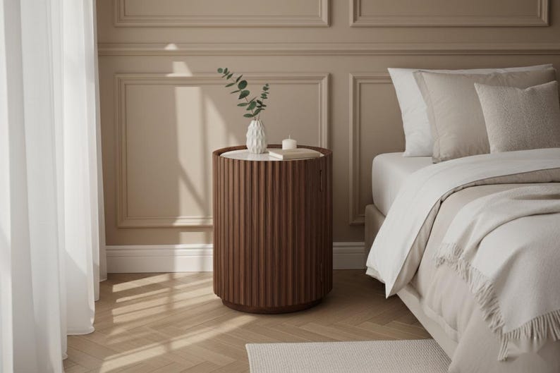 May include: A brown, fluted side table with a white top, holding a white vase with greenery and a small candle. The table is next to a bed with white bedding and pillows. The room has beige walls and a light wood floor.