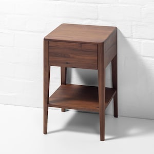 Against a white brick wall backdrop, an angled front-right view shows a handmade American walnut nightstand, a compact, solid wood bedside table with rich, warm walnut wood grain and sleek, curved corners. This modern hardwood furniture piece features a soft close drawer and an open lower shelf, highlighting expert craftsmanship, a hand-finished surface, and a minimalist design.