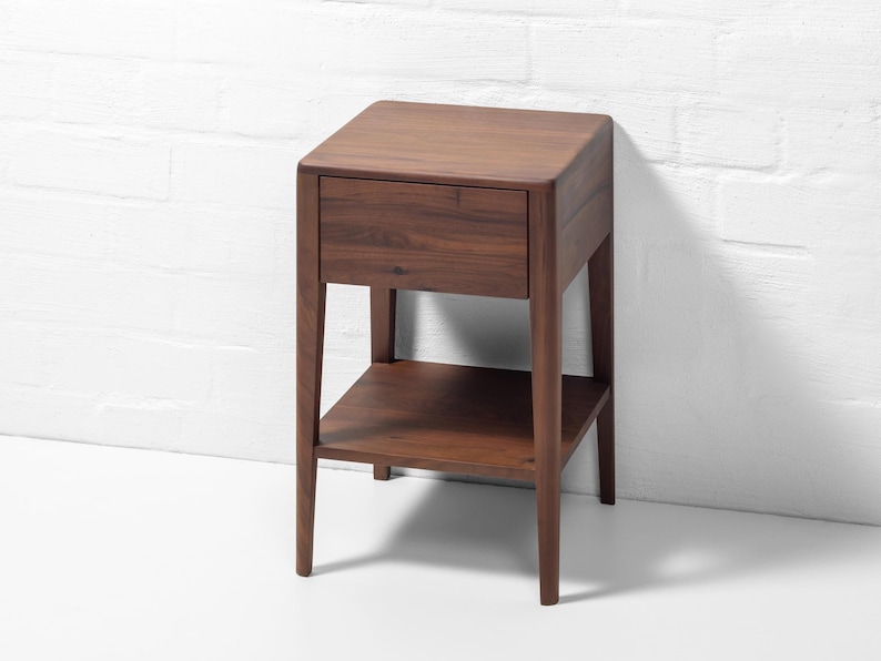 Against a white brick wall backdrop, an angled front-right view shows a handmade American walnut nightstand, a compact, solid wood bedside table with rich, warm walnut wood grain and sleek, curved corners. This modern hardwood furniture piece features a soft close drawer and an open lower shelf, highlighting expert craftsmanship, a hand-finished surface, and a minimalist design.