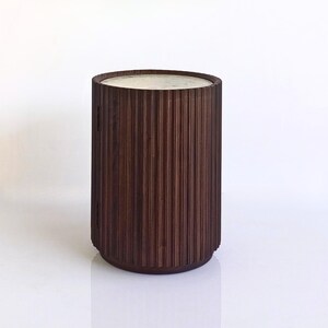 May include: A dark brown, cylindrical side table with a light-colored top. The table has vertical, ridged detailing around the sides. The design is modern and minimalist, suitable for various interior styles.