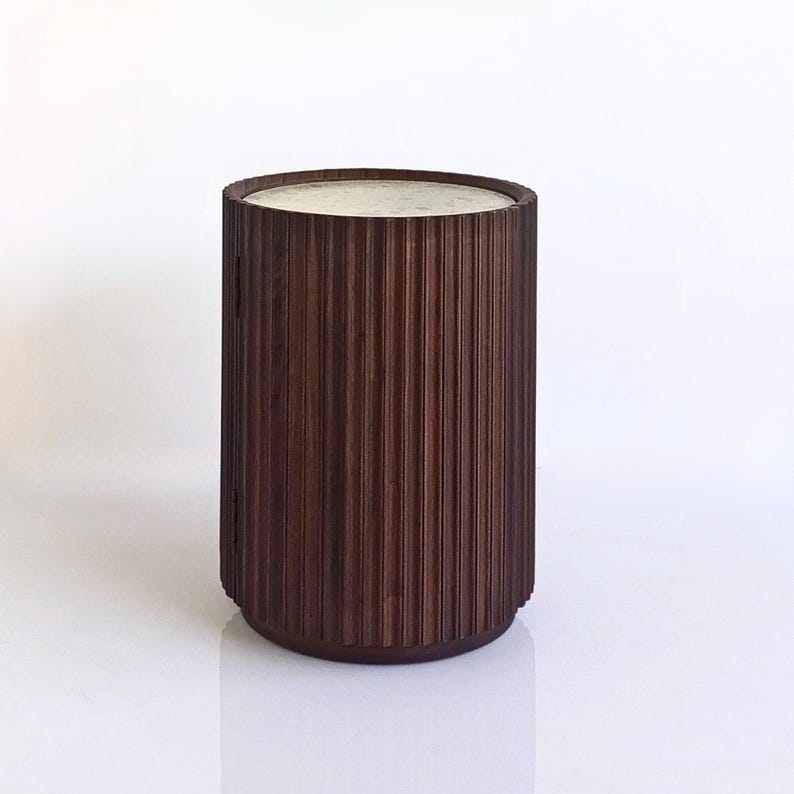 May include: A dark brown, cylindrical side table with a light-colored top. The table has vertical, ridged detailing around the sides. The design is modern and minimalist, suitable for various interior styles.