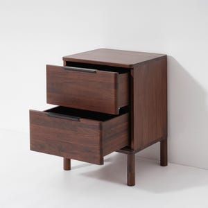 May include: A dark brown wooden nightstand with two drawers, both partially open. The nightstand has a rectangular shape with a flat top and sits on four short legs. The drawer pulls are a dark color, contrasting with the wood grain.