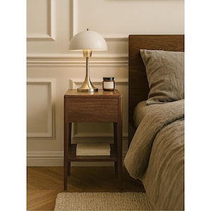 May include: A mid-century modern style bedroom scene featuring a wooden nightstand with a drawer and shelf. A white dome-shaped lamp with a brass base sits on top, next to a small jar. A bed with a linen duvet is partially visible.