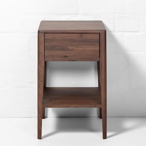 A handmade American walnut nightstand stands centered in front of a white brick wall backdrop, a compact, modern wood bedside table featuring a closed soft-close drawer and an empty lower shelf. This solid walnut furniture piece showcases a rich natural wood grain, warm brown tone, smooth finish, and clean silhouette, with the white brick backdrop emphasizing its minimalist modern design.