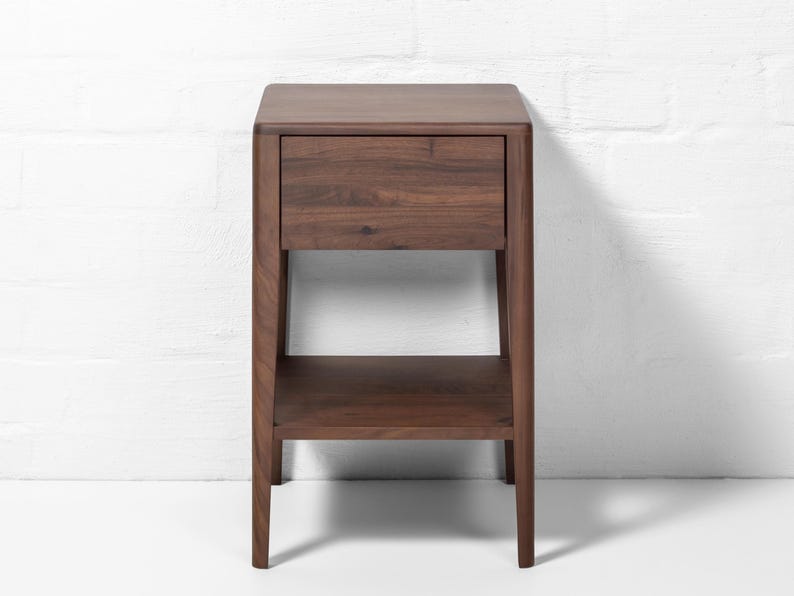 A handmade American walnut nightstand stands centered in front of a white brick wall backdrop, a compact, modern wood bedside table featuring a closed soft-close drawer and an empty lower shelf. This solid walnut furniture piece showcases a rich natural wood grain, warm brown tone, smooth finish, and clean silhouette, with the white brick backdrop emphasizing its minimalist modern design.