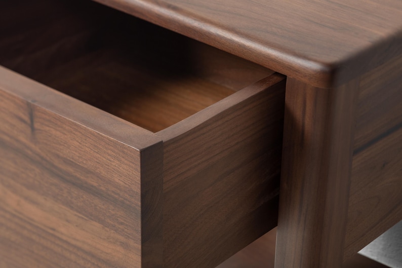A close-up of the corner of a luxury, heirloom-quality American walnut nightstand with the soft-close drawer open, showcasing meticulous craftsmanship. Rich walnut wood grain and smooth hand-finished surfaces highlight fine detailing and expert joinery, reflecting artisanal craftsmanship in this handcrafted furniture piece. Clean lines and tight-fitting joints emphasize fine workmanship in this compact bedside table.