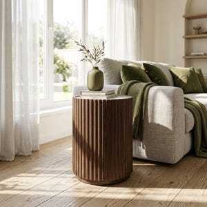 May include: A cylindrical side table with a dark brown wood finish and a vertically fluted, ribbed exterior. The table features a flat top surface holding two stacked books and a matte olive green vase containing a small leafy branch.