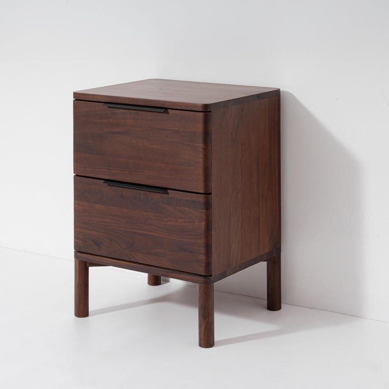 May include: A dark brown wooden nightstand with two drawers and black handles. The nightstand has a rectangular shape with rounded edges and sits on four short, cylindrical legs. The wood grain is visible.