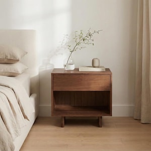 May include: A wooden nightstand with a drawer and open shelf, next to a bed with beige bedding. A small vase with greenery, a book, and a decorative object sit on top of the nightstand. The room has a neutral color palette.