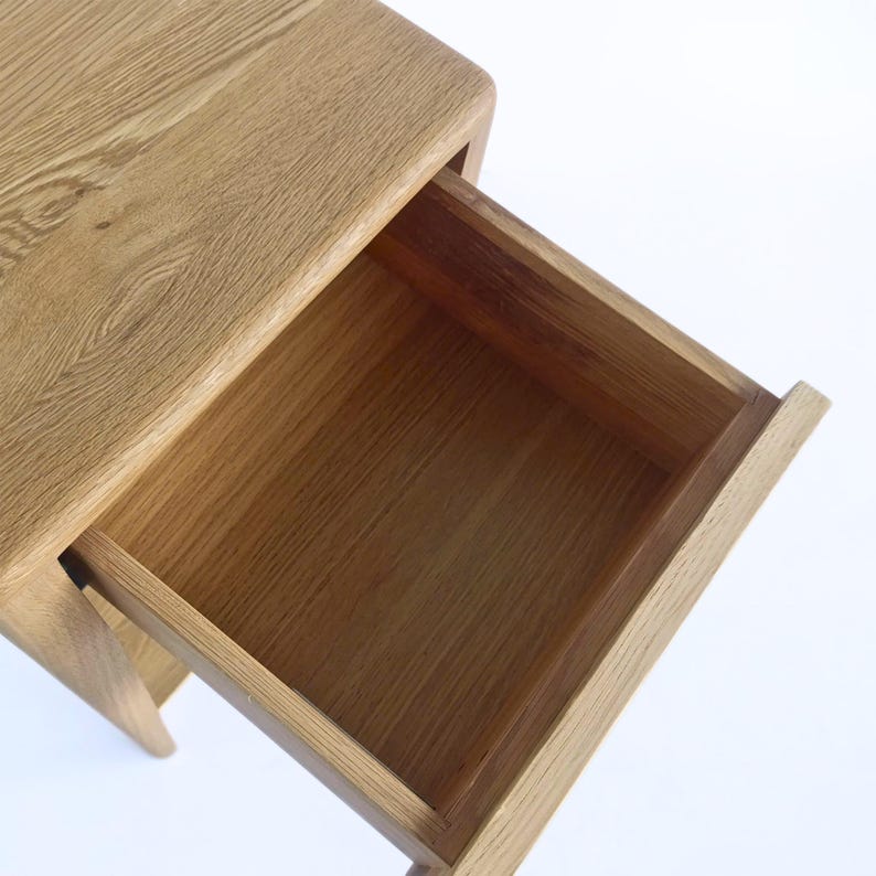 Close-up angled view of a handmade American white oak nightstand, showing the open drawer and interior craftsmanship. The Scandinavian bedside table highlights smooth wood grain, natural light oak finish, and clean lines with softly rounded edges. Made from solid hardwood, this modern oak furniture piece emphasizes minimalist design and durable handmade construction.
