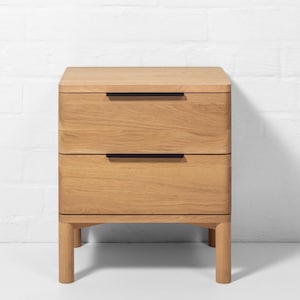 May include: A light oak two-drawer nightstand with black handles. The nightstand has a rounded design and sits on four short legs. The wood grain is visible on the drawers and top. The background is a white brick wall.
