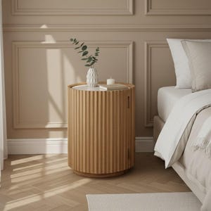 May include: A light brown, ribbed wooden bedside table with a round top. A white vase with greenery and a small candle sit on top. The table is next to a bed with white bedding and pillows.