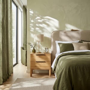 May include: A light oak nightstand with two drawers and black handles, topped with a beige lamp, a small green vase with a plant, and a silver alarm clock. The nightstand sits beside a bed with olive green bedding and a beige upholstered headboard.