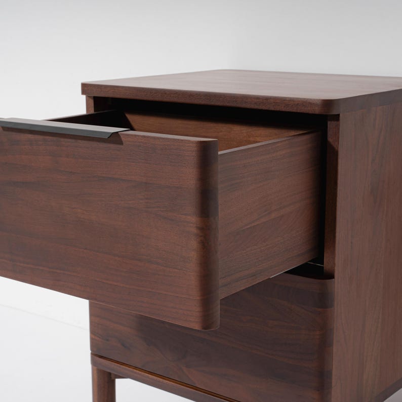 May include: A dark brown wooden nightstand with a drawer partially open, revealing the interior. The nightstand has a rectangular shape with rounded edges and a dark metal handle. The wood grain is visible on the surface.