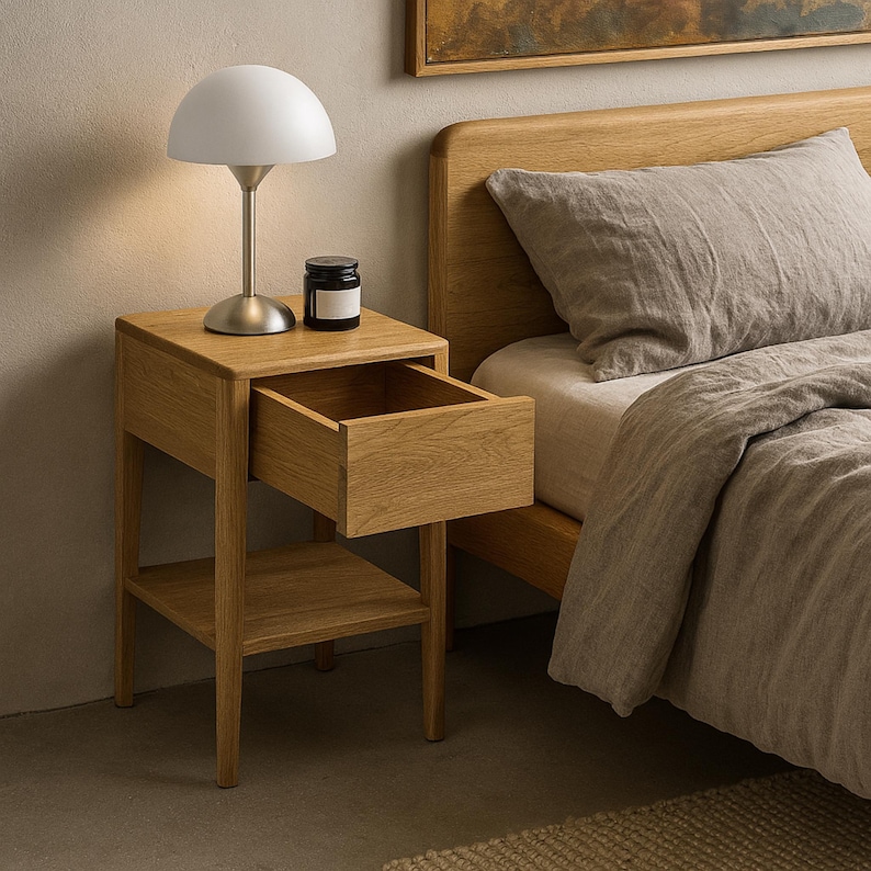In a styled neutral bedroom, a small Scandinavian oak nightstand, handcrafted from American White Oak, stands beside a wood-framed bed. The small wood bedside table with drawer has its drawer fully open, revealing the wood interior, and an empty lower shelf. A white dome lamp with a brushed base and a dark candle jar sit on top, highlighting the modern handcrafted nightstand’s minimalist style.