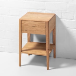 Handmade American white oak nightstand photographed from a front-right angle against a white brick wall. This small Scandinavian bedside table features a single handle-free soft-close drawer and a lower open shelf. The solid oak hardwood showcases natural grain, smooth finish, and tapered legs, emphasizing minimalist craftsmanship and modern functional design for bedroom storage.