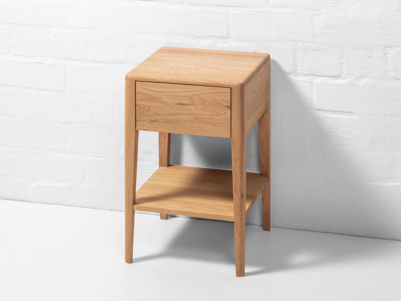 Handmade American white oak nightstand photographed from a front-right angle against a white brick wall. This small Scandinavian bedside table features a single handle-free soft-close drawer and a lower open shelf. The solid oak hardwood showcases natural grain, smooth finish, and tapered legs, emphasizing minimalist craftsmanship and modern functional design for bedroom storage.