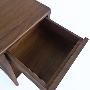 Overhead close-up of a handmade walnut nightstand with its drawer fully open, focusing on the wood drawer detail and fine craftsmanship. This solid wood bedside table’s drawer reveals precise dovetail joinery, smooth sanded interior surfaces, and ample storage space. Rich American walnut wood grain patterns and gently curved edges showcase expert woodworking and quality construction.