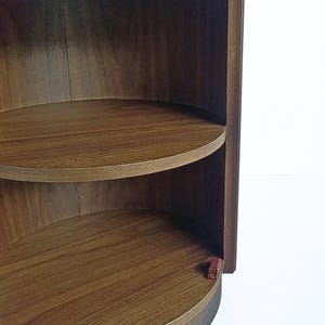 May include: A dark brown, semi-circular wooden shelf unit with two shelves. The shelves are curved and the wood grain is visible. The unit is set against a white background.