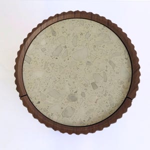 May include: A round serving tray with a light-colored terrazzo surface and a dark brown, scalloped wooden rim. The tray's diameter is approximately 12 inches.