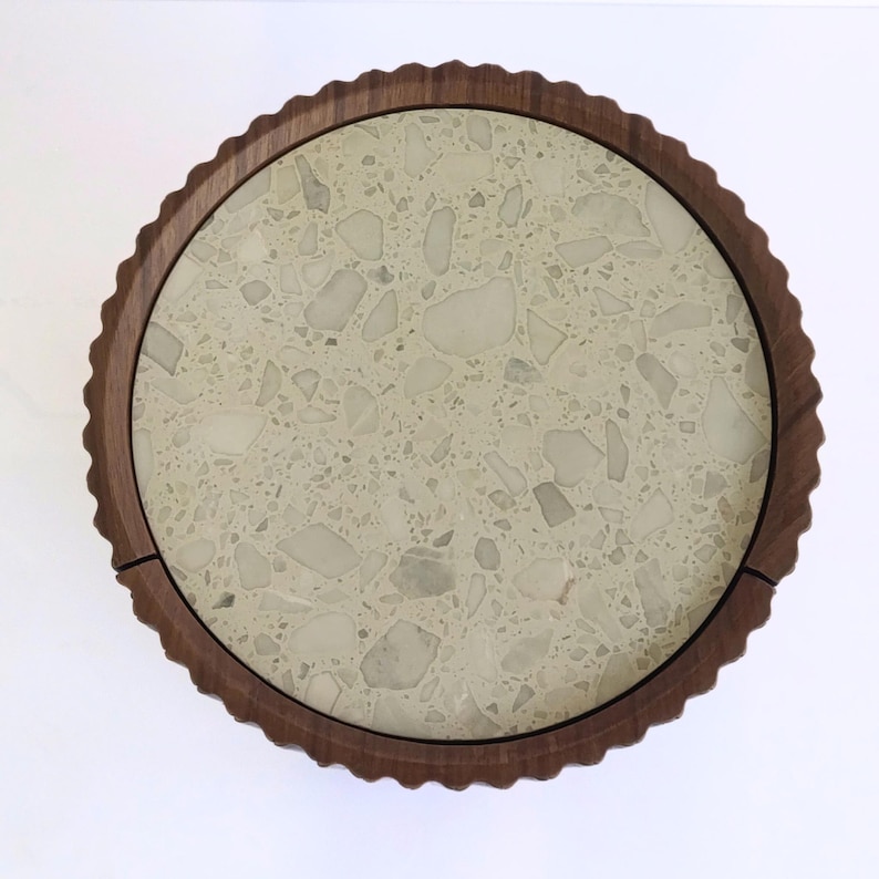 May include: A round serving tray with a light-colored terrazzo surface and a dark brown, scalloped wooden rim. The tray's diameter is approximately 12 inches.