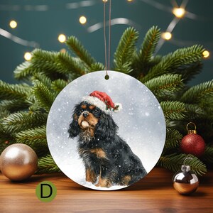 May include: Round Christmas ornament featuring a Cavalier King Charles Spaniel wearing a Santa hat. The dog is black and tan, set against a snowy background. The ornament has a string for hanging and is surrounded by Christmas tree branches.