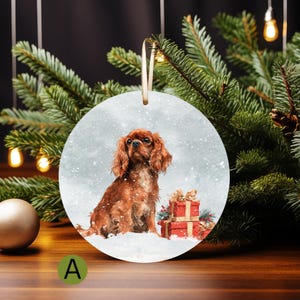 May include: A round Christmas ornament featuring a Cavalier King Charles Spaniel dog sitting in the snow next to two red gift boxes. The ornament has a beige ribbon for hanging and is surrounded by a Christmas tree.