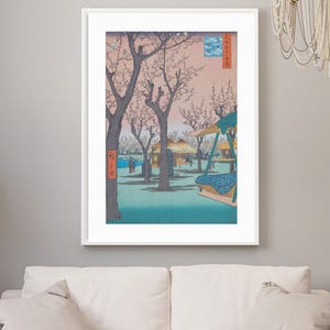 May include: A framed print of a Japanese landscape scene. The artwork depicts a park with cherry blossom trees, figures, and traditional buildings. The colour palette includes pink, blue, and brown tones. The print is displayed above a white sofa.