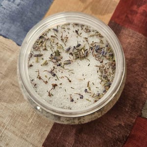 May include: A clear plastic jar filled with white bath salts and dried lavender and herbs. The jar is round with a clear lid. The background is a multi-colored fabric with shades of blue, brown, and red.