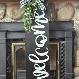May include: A black wooden welcome sign with white cursive lettering. The sign is decorated with a black and white checkered bow, greenery, and small white flowers. The sign is displayed in front of a fireplace.
