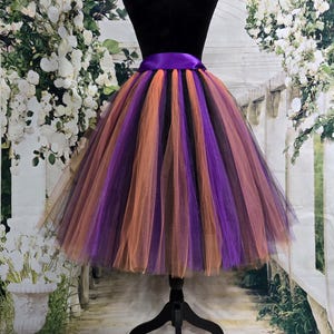 Könnte beinhalten: Ein mehrlagiger Tüllrock in Lila, Orange und Schwarz mit einem breiten, violetten Satinbund. Der Rock wird auf einer schwarzen Schaufensterpuppe präsentiert, um seine volle, ausgestellte Form zu zeigen. Geeignet für Kostüme oder besondere Anlässe.