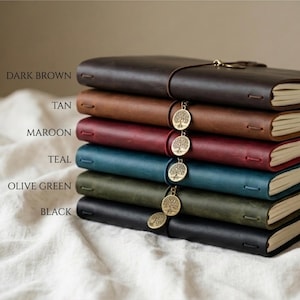Puede incluir: Una pila de seis diarios de cuero en varios colores: marrón oscuro, marrón claro, granate, verde azulado, verde oliva y negro. Cada diario está encuadernado con una correa de cuero y tiene un dije dorado con un diseño de árbol.