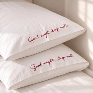 May include: Two white pillowcases stacked on top of each other. Each pillowcase features the embroidered text "Good night, sleep well" in a dark red cursive font across the front panel, with clean stitching and a smooth fabric finish.