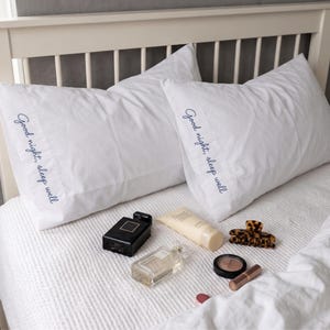 May include: Two white pillowcases featuring the embroidered navy blue text "Good night, sleep well" on the side. The pillows are arranged on a bed alongside a black perfume bottle, a clear glass perfume bottle, a beige tube, a hair clip, and makeup.