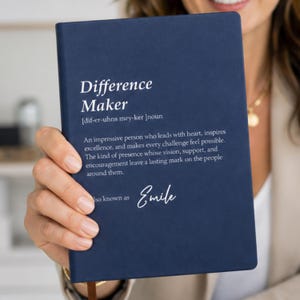以下が含まれることがあります： ネイビーブルーのノートブック。白い文字で「Difference Maker [dif-er-uhns mey-ker] noun. An impressive person who leads with heart, inspires excellence, and makes every challenge feel possible. Also known as Emile.」と書かれています。