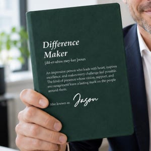 Può includere: Un taccuino verde scuro con testo bianco che definisce il termine Difference Maker come una persona impressionante che guida con il cuore e ispira eccellenza. Sotto la definizione, il testo recita Also known as Jason in corsivo bianco.