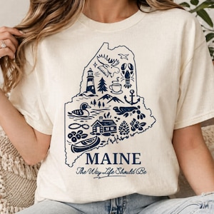 Könnte beinhalten: Ein cremefarbenes Kurzarm-T-Shirt mit einer dunkelblauen Grafik des Bundesstaates Maine. Das Design enthält Illustrationen eines Elchs, Hummers, Leuchtturms, einer Hütte, eines Kanus und anderer Symbole sowie den Text MAINE The Way Life Should Be.