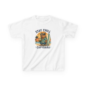 Op de afbeelding: Wit T-shirt met een cartoon capibara met een zonnebril en een Hawaiiaans shirt, met een tropisch drankje en leunend tegen een surfplank. De tekst "STAY CHILL CAPYBARA" staat boven en onder de illustratie.
