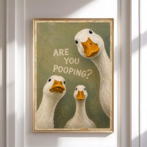 Può includere: Una stampa artistica da parete incorniciata raffigurante tre anatre bianche con becchi arancioni su uno sfondo verde salvia testurizzato. Il testo ARE YOU POOPING? è stampato in color crema chiaro al centro sopra le due anatre più piccole.
