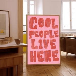 Op de afbeelding: Een ingelijste roze poster met de woorden "COOL PEOPLE LIVE HERE" in rode, vette letters. De poster wordt weergegeven op een houten vloer, leunend tegen een muur. Het frame heeft een lichtbruine kleur.
