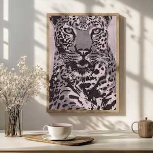 May include: A framed print of a leopard's face in black and white tones. The artwork is displayed on a white surface, with a cup of coffee, a wooden tray, and a vase of white flowers in the foreground. The frame is a light brown color.